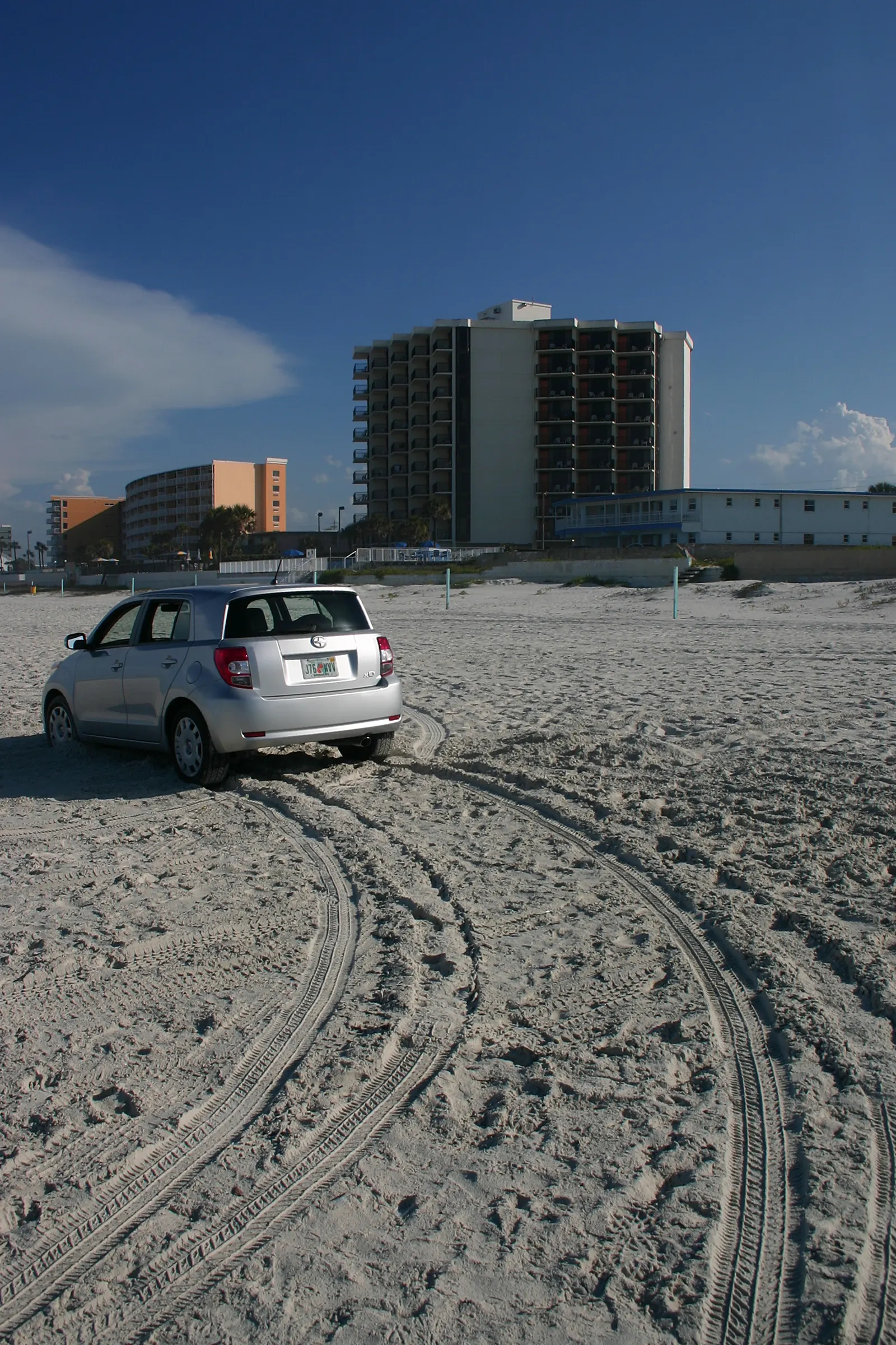 Silver hatchback on packed beach sand with tire tracks and beachfront buildings behind