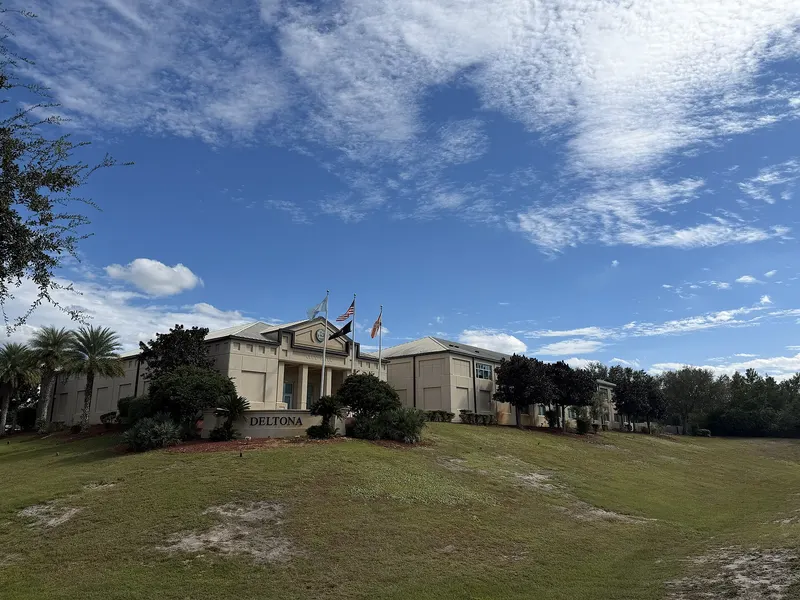 Deltona City Hall on a grassy rise with flags, palms and a DELTONA monument sign under a blue sky