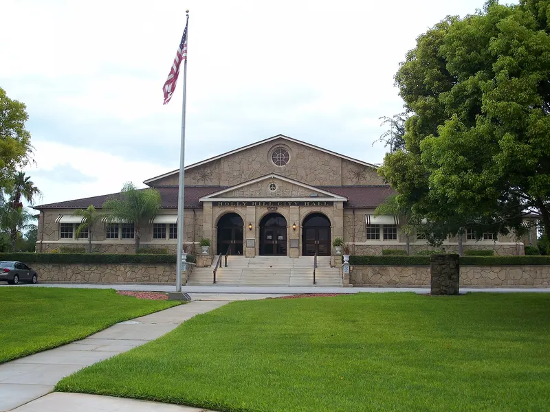 Stone-faced Holly Hill City Hall with three arched entrances, front steps, a flagpole and a green lawn