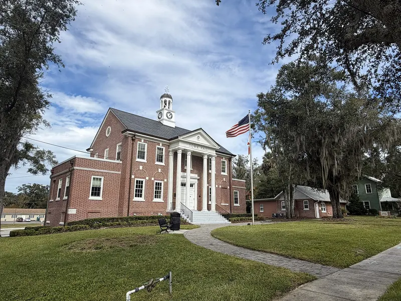 Red-brick Orange City Town Hall with white columns and a clock cupola, flag and moss-draped oaks
